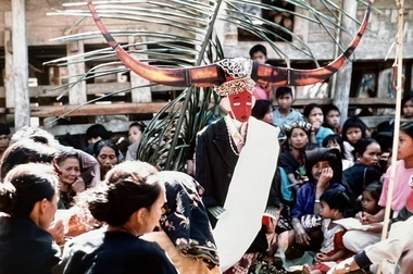 Tau-tau funerary effigy with a white sarong and a red face, surrounded by women, at Pangleon (Salupputti), 1993. The horns indicate the deceased is male., Effigie funéraire tau-tau, entourée de femmes, à Pangleon (Saluputti), 1993. Les cornes indiquent que le défunt est masculin. (French), Patung tau-tau dikerumuni oleh kaum perempuan, di Pangleon (Saluputti), 1993. Tanduk itu merupakan tanda bahwa mendiang adalah seorang lelaki. Patung itu mengenakan sarung putih, dengan wajah yang berwarna merah. (Indonesian) thumbnail